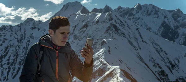 A young man with phone in hand on the top of a snowy mountain far from civilization on a background of blue sky. The concept of activity and the availability of mobile connection