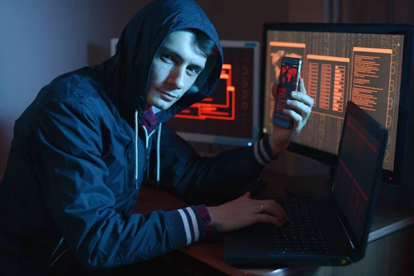 Male hacker in the hood holding the phone in his hands trying to hack the mobile device cloud and steal data in neon light. The concept of cyber security