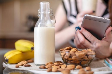 Mutfakta elinde bir telefon tutan organik badem sütü içen kadın. Şişe ve meyve masada. Diyet vejetaryen ürün