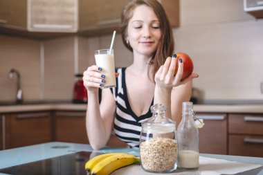 Mutfakta cam bardak tutan pipetlerden organik yulaf sütü içen kadın. Şişe ve meyveler masada. Diyet sağlıklı vejetaryen ürün