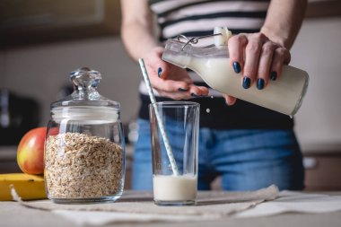 Bir kadın mutfakta ki masanın üzerindeki bardağa şişeden organik yulaf sütü döker. Diyet sağlıklı vejetaryen ürün