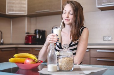 Mutfakta cam bardak tutan pipetlerden organik yulaf sütü içen kadın. Şişe ve meyveler masada. Diyet sağlıklı vejetaryen ürün