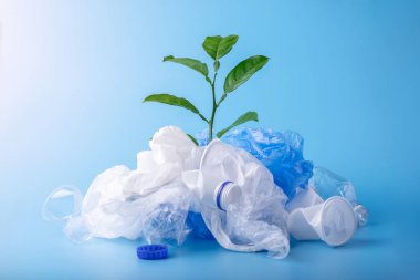 Green plant grows among plastic garbage. Bottles and bags on blue background. Concept of environmental protection and waste sorting. Place for text clipart