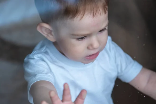 Lindo niño mirando en el cristal de la ventana con reflejo. Soledad de los niños y espera de la bondad . — Foto de Stock