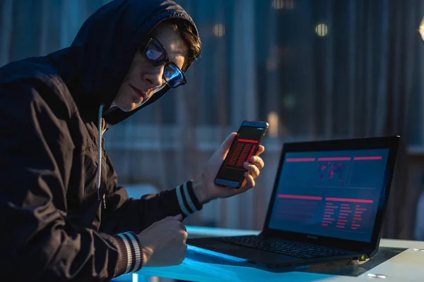 Pria hacker di kap memegang telepon di tangannya mencoba untuk mencuri akses database. Konsep keamanan dunia maya — Stok Foto