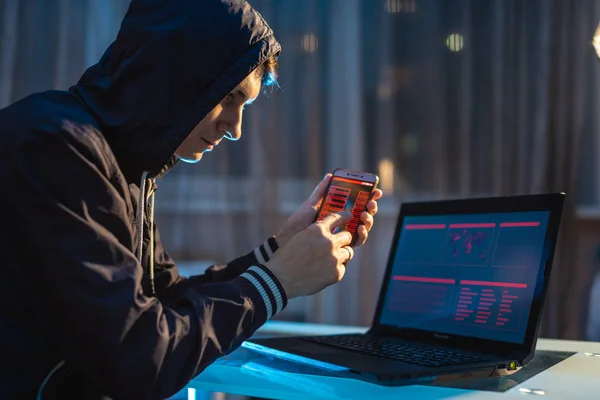 Pria hacker di kap memegang telepon di tangannya mencoba untuk mencuri akses database. Konsep keamanan dunia maya — Stok Foto