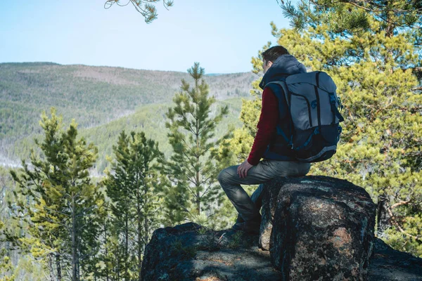 Hombre excursionista con una gran mochila sentado en un acantilado de roca y mirando el extenso valle verde. Libertad en los viajes — Foto de Stock