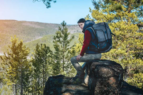 Man hiker with a large backpack sitting on a rock cliff and looking at the sprawling green valley. Freedom in travel — Stock Photo, Image