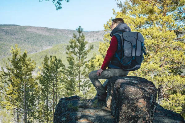 Hombre excursionista con una gran mochila sentado en un acantilado de roca y mirando el extenso valle verde. Libertad en los viajes — Foto de Stock