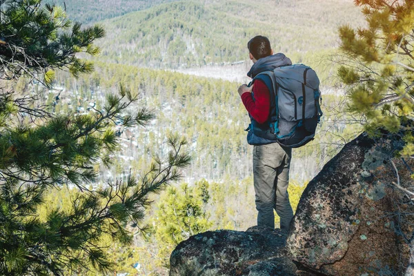 Man tourist with a large backpack stands on a rock cliff and looking at the sprawling green valley. Freedom in travel — Stock Photo, Image