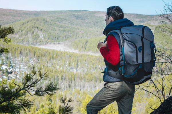 Homem turista com uma grande mochila fica em um penhasco de rocha e olhando para o vale verde alastrando. Liberdade de circulação — Fotografia de Stock