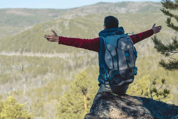 L'escursionista uomo con un grande zaino si erge su una scogliera di pietra di fronte a una valle verde con le braccia tese — Foto Stock