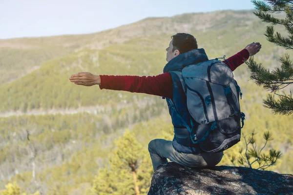 Homem caminhante com uma grande mochila fica em uma pedra penhasco na frente de um vale verde com os braços estendidos — Fotografia de Stock