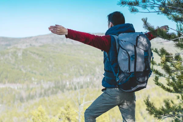 Homem caminhante com uma grande mochila fica em uma pedra penhasco na frente de um vale verde com os braços estendidos — Fotografia de Stock