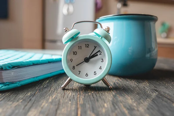 Μπλε ρολόι ξυπνητήρι στο τραπέζι με μια κούπα και ένα ημερολόγιο στο φόντο της κουζίνας. Λειτουργία ώρας και οικίας — Φωτογραφία Αρχείου