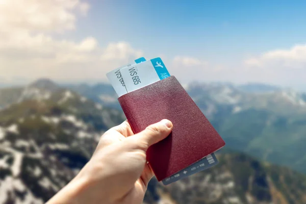 Kezében útlevél és két repülőjegyet a kék háttérben a hegyek. Utazás és nyaralás gyönyörű helyeken — Stock Fotó