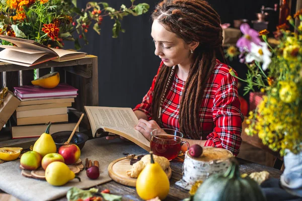 Belle jeune femme lisant un livre romantique dans une atmosphère automnale. Table rustique avec des fleurs jaunes et une pile de livres — Photo