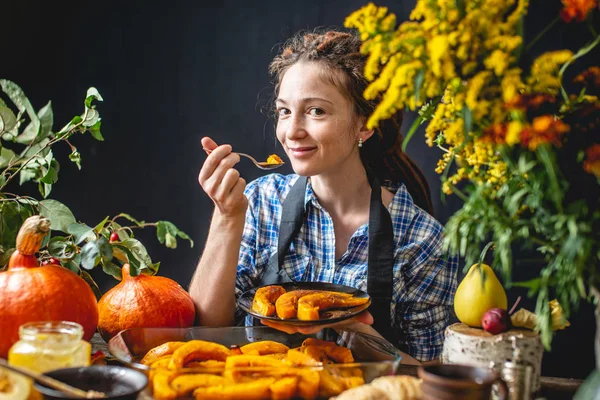 Mujer chef sosteniendo rebanadas de calabaza de naranja al horno con miel y canela. Comida de otoño en una acogedora cocina con flores . — Foto de Stock