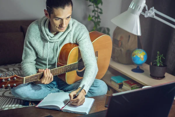 Young male musician is learning to play acoustic guitar in an online lesson using laptop, at night by the light of a lamp in a room at home..