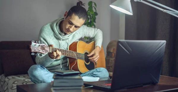 Genç erkek müzisyen, evdeki bir odada gece lamba ışığında dizüstü bilgisayar kullanarak online bir derste akustik gitar çalmayı öğreniyor...