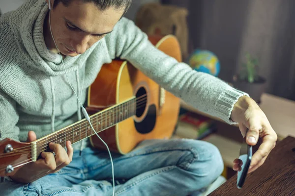 一位年轻的音乐家正在学习如何用电话应用程序在网上的一节课上弹奏吉他 一个男人在家里的一个房间里借着一盏灯学习功课 — 图库照片