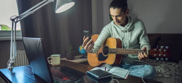 A young musician is learning to play acoustic guitar in an online lesson using a phone app. A man is studying courses at home in a room by the light of a lamp