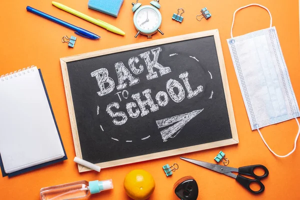 Stationery school supplies, medical mask and antiseptic around the board with the words Back to school. Orange background, top view.