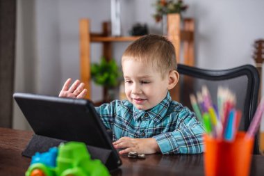 Gömlekli küçük çocuk tablet ekranına bakıyor ve el sallıyor. Çevrimiçi iletişim ve uzaklık eğitimi kavramı