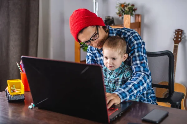 Young modern man father is working on a laptop, and his little son is sitting on his lap. Concept of family and remote work from home