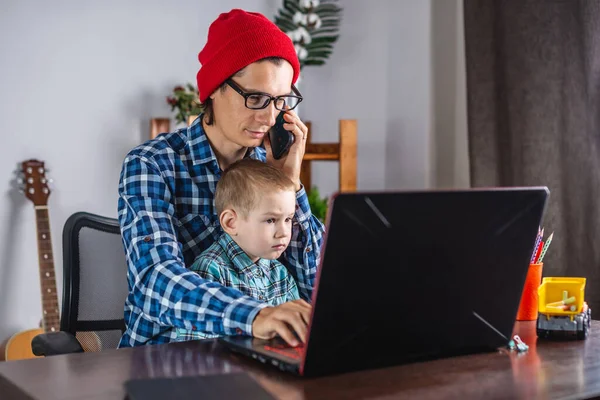 Young modern man father is working on a laptop, and his little son is sitting on his lap. Concept of family and remote work from home
