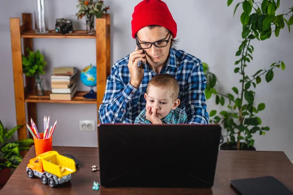 Young modern man father is working on a laptop, and his little son is sitting on his lap. Concept of family and remote work from home