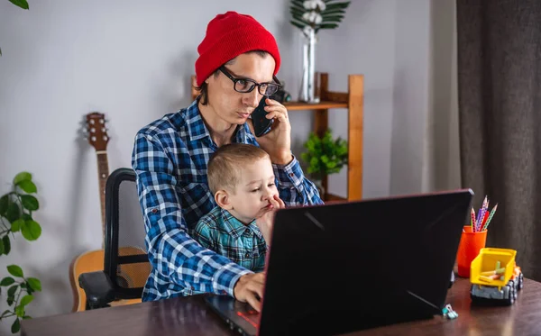 若い現代人の父はノートパソコンで働いていて 彼の小さな息子は彼の膝の上に座っています 家庭からの仕事とリモートワークの概念 — ストック写真