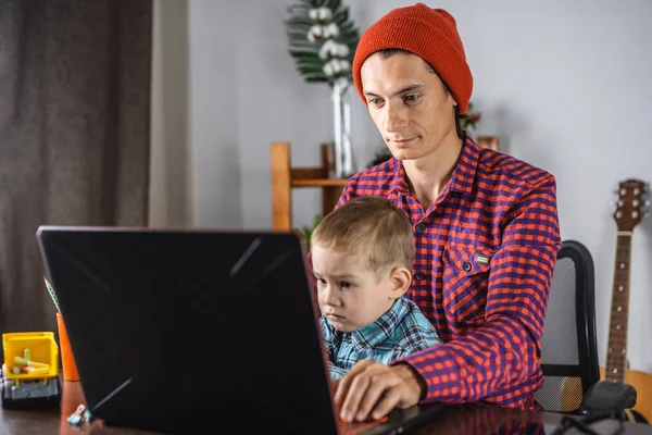 Young modern man father is working on a laptop, and his little son is sitting on his lap. Concept of family and remote work from home