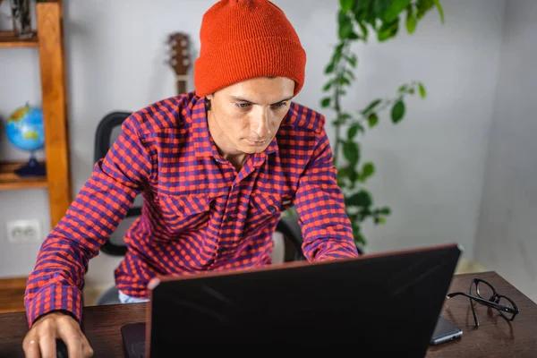 Modern young man in a red checked shirt and hat is working on a laptop. Concept of distance communication and remote job.