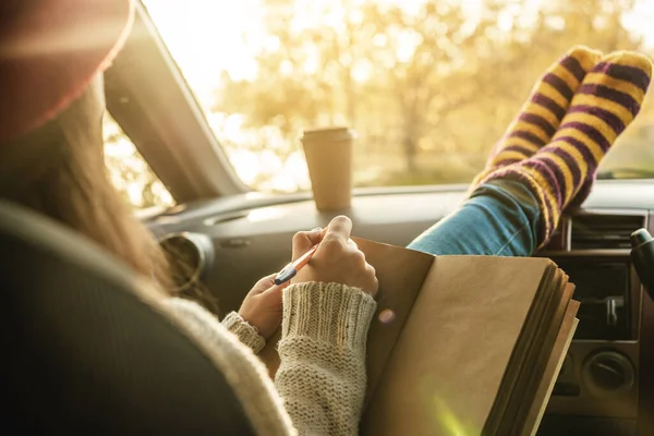 Woman in a car in warm socks is writing notes and plans in a notebook. Cozy autumn weekend trip. The concept of freedom of travel