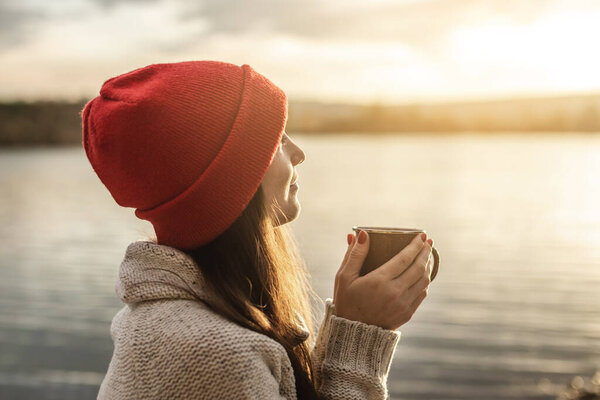Woman in a red hat is drinking coffee on the lake at sunset. Autumn outdoor recreation. The concept of freedom of travel