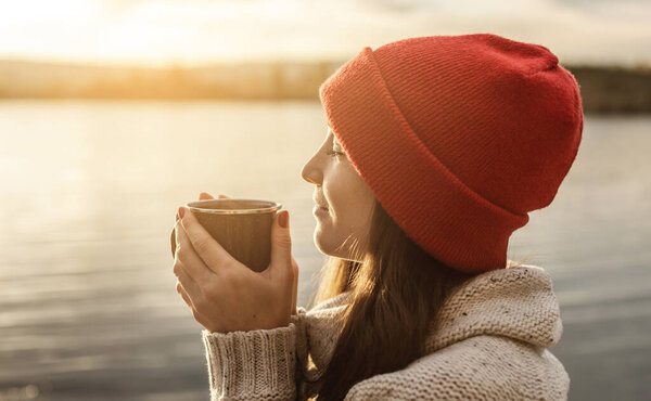 Woman in a red hat is drinking coffee on the lake at sunset. Autumn outdoor recreation. The concept of freedom of travel