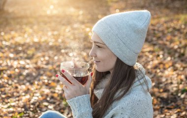 Genç ve güzel bir kadın, sonbahar sarı parkında gün batımının ışığında sıcak çay içiyor. Soğuk sonbahar mevsiminde rahat bir yaşam tarzı kavramı
