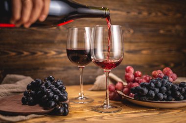 Bir el, bir şişeden bardağa kuru kırmızı şarap dökerken bir grup üzümün arka planına karşı ahşap bir masada. Eklem ve şarap üretimi kavramı