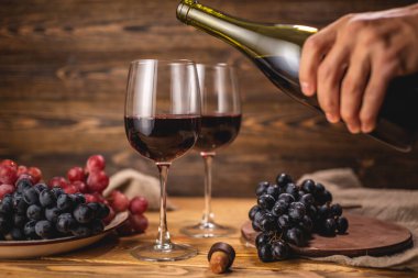 Bir el, bir şişeden bardağa kuru kırmızı şarap dökerken bir grup üzümün arka planına karşı ahşap bir masada. Eklem ve şarap üretimi kavramı