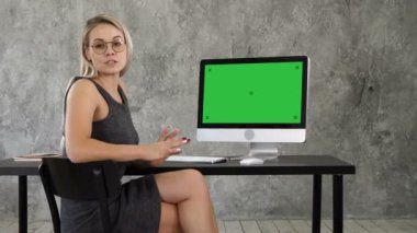 Kameraya konuşurken ve bilgisayar ekranında bir şey gösterilen ofisinde oturan neşeli genç kadın. Yeşil perde Mock-up göstermek.