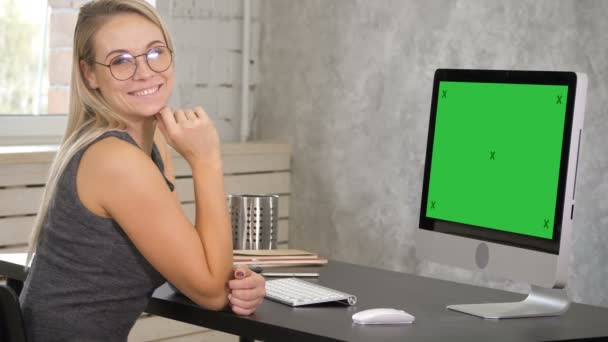 年轻女子微笑着对着坐在工作场所的相机。绿色屏幕模拟显示. — 图库视频影像