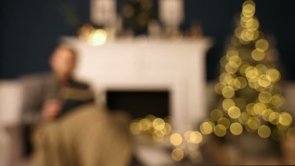 Un hombre con gafas en silla leyendo un libro en casa en Navidad en voz alta. Intérprete de Navidad . — Vídeos de Stock