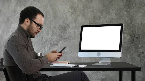 Uomo d'affari guardando la sua messaggistica smartphone e seduto vicino allo schermo del computer. Display bianco . — Foto Stock