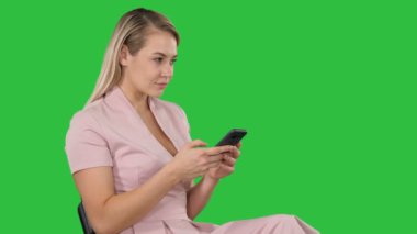 Oturan sarışın kadın gülümseyerek ve Chroma anahtar yeşil ekran smartphone kullanarak.