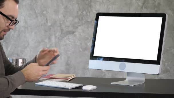 Un jeune homme envoie un texto au téléphone avec un ordinateur sur le bureau. Affichage blanc . — Video