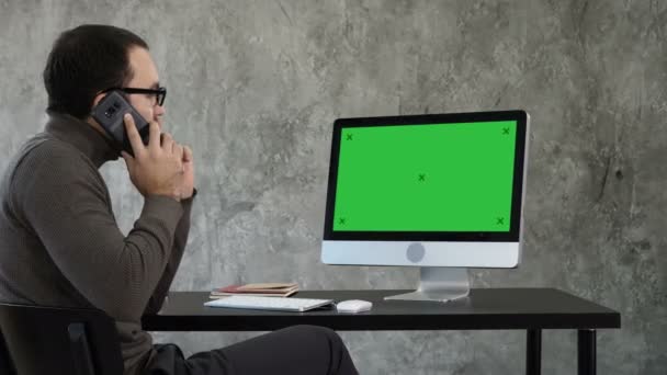Bilgisayarda çağdaş ofiste çalışan erkek tasarımcı. Telefon ve ekranda ne arıyorsunuz konuşuyor. Yeşil perde Mock-up göstermek. — Stok video