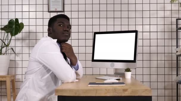 男性ハンサムのアフリカの医師は彼のコンピューターの隣に考えて。白ディスプレイ. — ストック動画