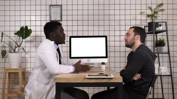 Doctor showing medical records on his computer to his patient, he is pointing at the screen. White Display. — Stock Video