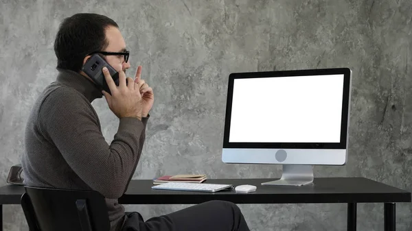 현대 사무실에 있는 컴퓨터에서 일 하는 남성 디자이너. 그는 휴대 전화에는 화면에 보고 이야기 한다. 화이트 디스플레이. — 스톡 사진